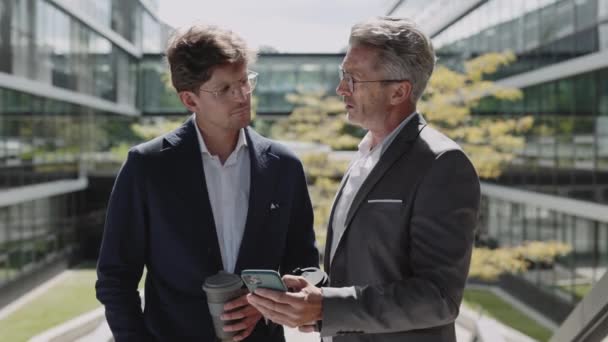 Dos hombres en traje de negocios usando móvil y hablando al aire libre — Vídeo de stock