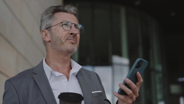 Hombre mayor en traje de pie al aire libre con café y móvil — Vídeo de stock