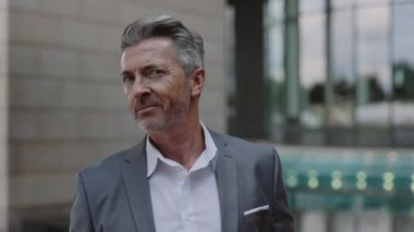 Gri takım elbiseli olgun bir adam şehrin sokaklarında kameraya gülümsüyor.