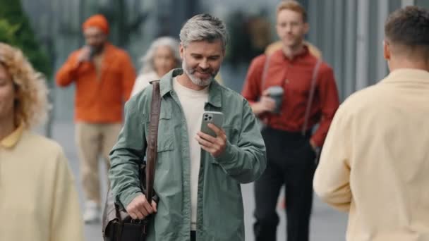 Smiling grey haired man walking in street and using mobile — Vídeos de Stock