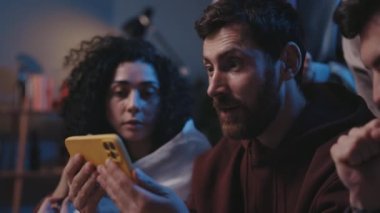 Akıllı telefondan maç sonuçlarını izlerken ve kanepede otururken çoklu etnik çeşitliliğe sahip insanların çığlıklarını kapat. Arkadaşlarıyla birlikte sevinen ve çığlık atan sakallı adama odaklan..