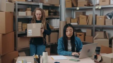 Rastalı kadının belindeki portre görüntüsü masaya paketler koyuyor ve Asyalı meslektaşına laptopta çalışırken bir şey gösteriyor. Küçük işletme kavramı.
