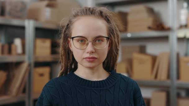 Портрет спокойной кавказской девушки с дредами в очках, смотрящей в камеру с серьезным лицом, стоя на рабочем месте с посылками на заднем плане. — стоковое видео