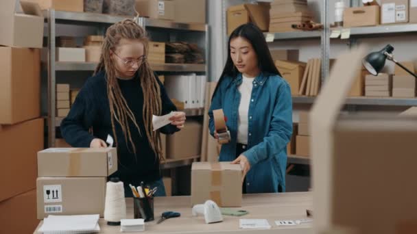 Two diverse female workers packing parcels with scotch tape while working together at the home office. Shelfs with cupboard boxes at the background. Small business concept. — Stock Video