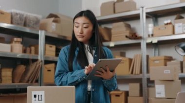 Tablet bilgisayarı kullanan Asyalı kadın bir şeyi incelerken ve müşteriye paket göndermeye hazırlanırken sakinleş. Depoda çalışan kız. Küçük işletme kavramı.