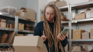Rastalı saçlı beyaz bir kadın akıllı telefon tutuyor ve paket hakkında bilgi hazırlarken klavyeye bir şeyler yazıyor. Posta servisi ve küçük işletme kavramı.
