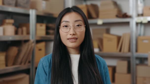 Портретний вигляд спокійна азіатська дівчина в окулярах дивиться на камеру з усміхненим обличчям, стоячи на її робочому місці з шафами на задньому плані . — стокове відео