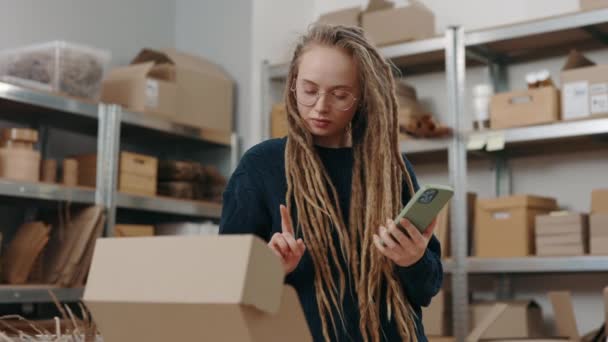 Vit kvinna med dreadlocks hålla smartphone och skriva något på tangentbordet samtidigt förbereda information om paket. Post service och småföretag koncept. — Stockvideo