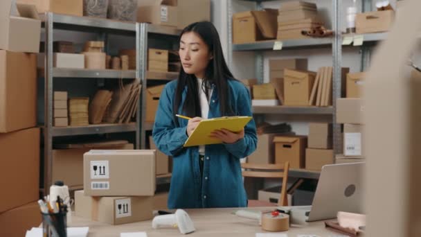 Waist up portrait view of the asian girl writing something at the folder while checking information about parcel and preparing sending it to the client. Small business concept. — Stock Video