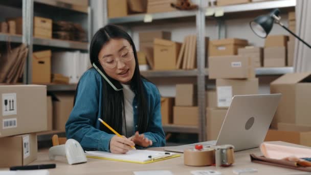 Taille-up portretweergave van de Aziatische vrouw chatten op de smartphone met de klant en het schrijven van pakketgegevens op de krant terwijl je achter de laptop. Post service en concept voor kleine bedrijven. — Stockvideo