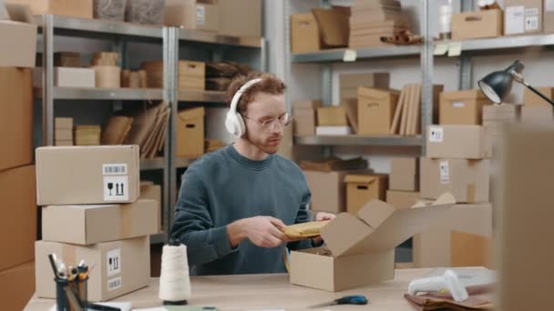 Figyelmes kaukázusi férfi szemüvegben és fülhallgatóval az asztalnál és csomagokat pakol az otthoni irodája szekrényében. Kisvállalkozói koncepció. — Stock videók