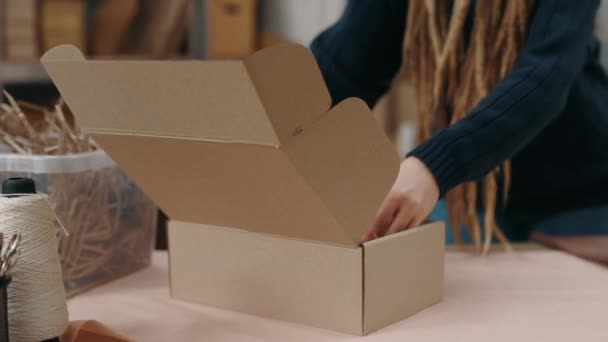 Närbild av den kaukasiska tonårskvinna med dreadlocks står vid lagret och packning paket vid skåpet lådor vid träbordet. Småföretagskoncept. — Stockvideo