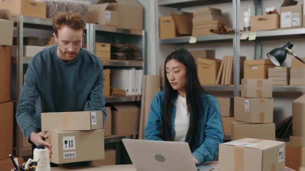Obraťte se na portrét bělocha, který pokládá balíčky na stůl a zkoumá je speciálním přístrojem poblíž svého asijského kolegy, zatímco ona pracuje u laptopu. Koncept malého podnikání. — Stock video