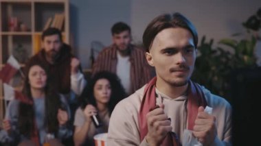 Heyecanlı bir beyaz erkek taraftar maç izlerken çığlık atıyor ve seviniyor ve arka planda arkadaşları olan favori takımın golünü kutluyor.