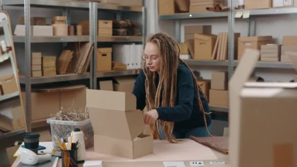 Attentive caucasian woman with dreadlocks wearing glasses standing at the table and packing parcels at the cupboard boxes at her home office. Small business concept. — Stock Video