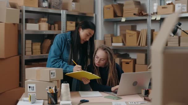 Aziatische brunette vrouw laat iets zien aan haar collega met een bril terwijl ze achter de laptop zit en iets typt op het toetsenbord. Concept voor kleine ondernemingen. — Stockvideo