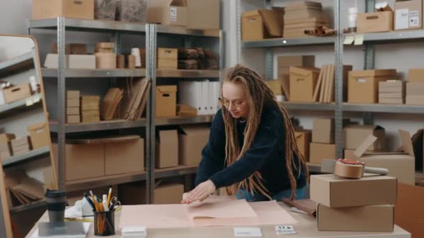 Werkproces. Portret van de zorgvuldige attente vrouw met dreadlocks op haar werkplek en pakjes met een lichte glimlach. Concept voor kleine ondernemingen. — Stockvideo