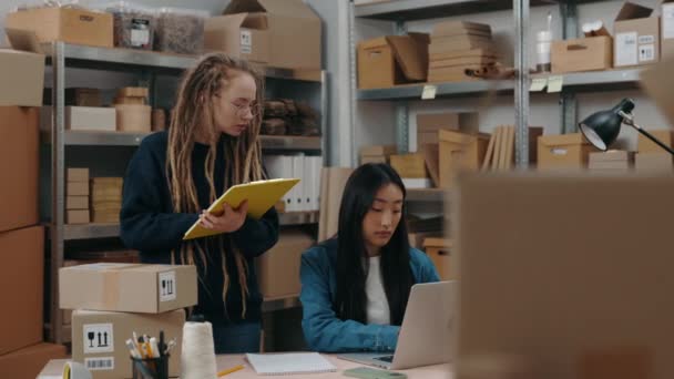 Confident woman with dreadlocks standing with folder at her hands and telling something to her asian colleague while she working at the laptop at office. Small business concept. — Stock Video