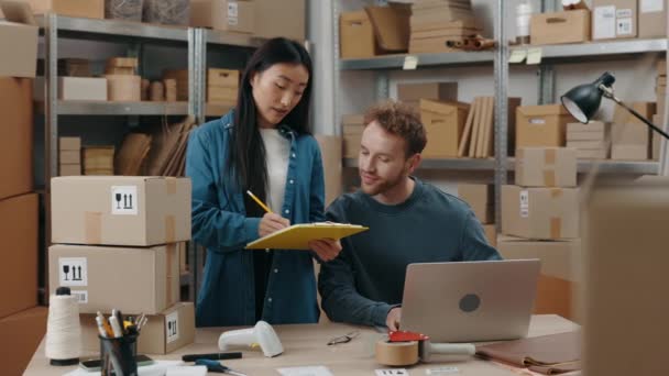 アジアのブルネットの女性は、彼がラップトップに座ってキーボードで何かを入力している間、眼鏡をかけて彼女の男性同僚に何かを示しています。中小企業コンセプト. — ストック動画