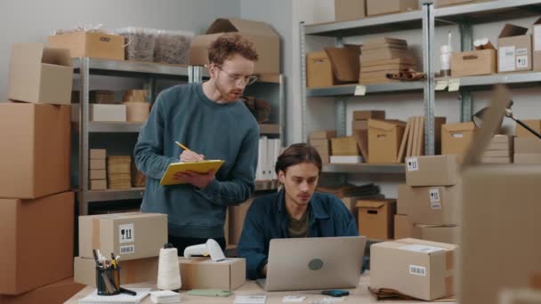 彼の男性の同僚が倉庫で彼と一緒に働いている間、ジンジャー白人男性はノートパソコンの画面を見て、紙にいくつかの詳細を書いています。中小企業コンセプト. — ストック動画