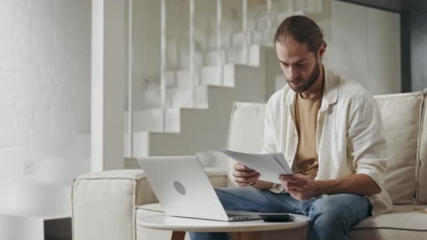 Hombre guapo haciendo papeleo mientras usa el portátil en casa — Vídeos de Stock