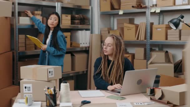 Skoncentrowana kobieta z dredami siedząca przy laptopie, podczas gdy jej azjatycka koleżanka stojąca z teczką przy półkach i pytająca o coś. Koncepcja małych przedsiębiorstw. — Wideo stockowe