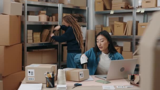 Asiatisk kvinna skanna skåp paket låda och skriva paket detaljer på laptop samtidigt berätta något för sin kollega. Flicka med dreadlocks sorterar igenom paket. Småföretagskoncept. — Stockvideo