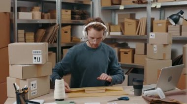 İşlem. Dikkatli, dikkatli, kulaklık takan, iş yerinde oturan ve müzik dinlerken hafif gülümsemeli paketler hazırlayan adamın portre görüntüsünü yükseltin. Küçük işletme kavramı.