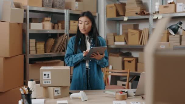 Widok z pasa w górę na azjatycką dziewczynę wpisującą coś na tablecie podczas sprawdzania informacji o paczce i przygotowywania wysyłania jej do klienta. Koncepcja małych przedsiębiorstw. — Wideo stockowe