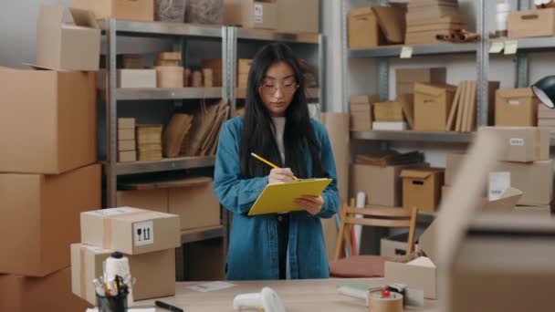 Spokojna Azjatka pisząca coś w folderze i przygotowująca przesyłkę do klienta. Dziewczyna pracująca w magazynie. Koncepcja małych przedsiębiorstw. — Wideo stockowe