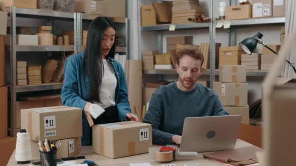 Vista de la cintura hacia arriba del retrato de la mujer asiática escaneando paquetes con un dispositivo especial en la mesa cerca de su colega masculino mientras él trabaja en el portátil. Concepto de pequeña empresa. — Vídeos de Stock