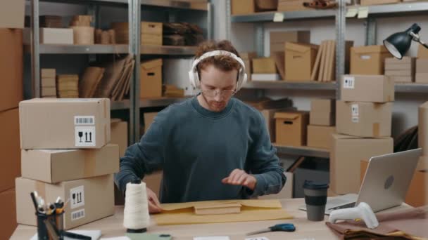 Procesu. Widok w pasie w górę portretu uważnego człowieka noszącego słuchawki siedzącego w miejscu pracy i pakującego paczki z lekkim uśmiechem podczas słuchania muzyki. Koncepcja małych przedsiębiorstw. — Wideo stockowe