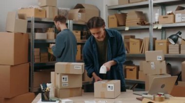 Esmer adam dizüstü bilgisayar ekranına bakıyor ve erkek arkadaşı depoda onunla çalışırken özel bir cihazla paketi tarıyor. Küçük işletme kavramı.