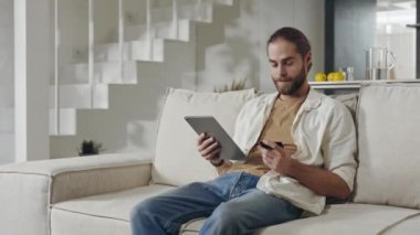 Tablet ve banka kartıyla koltukta oturan adam.