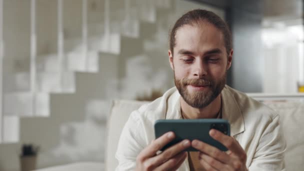 Homem barbudo assistindo vídeo no smartphone enquanto sentado no sofá — Vídeo de Stock