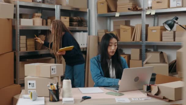 Femme caucasienne concentrée debout avec le dossier à ses mains près des étagères et demandant à son collègue asiatique pendant qu'elle travaille à l'ordinateur portable au bureau. Concept de petite entreprise. — Video