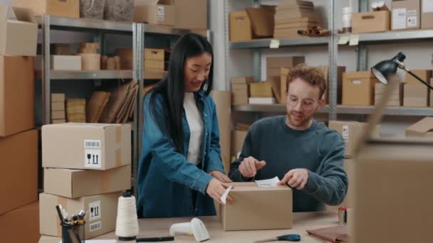 Mladí majitelé mužského a ženského internetového obchodu uzavírají balíčky lepicí páskou, zatímco připravují doručovací balíček ve skladu. Poštovní služba a koncept malého podnikání. — Stock video