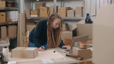 Masada duran beyaz bir kadın dizüstü bilgisayara bakıyor ve depoda çalışırken paket teslimatı hakkında bilgi yazıyor. Posta servisi ve küçük işletme kavramı.