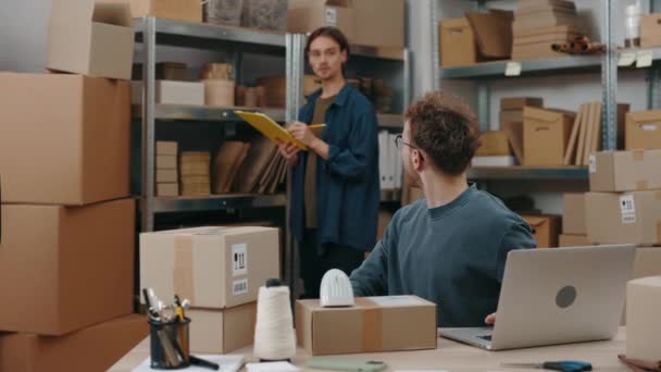 Ingefära kaukasier man tittar på sin manliga kollega medan du arbetar på lagret och skriva paketuppgifter vid laptop datorn. Post service och småföretag koncept. — Stockvideo