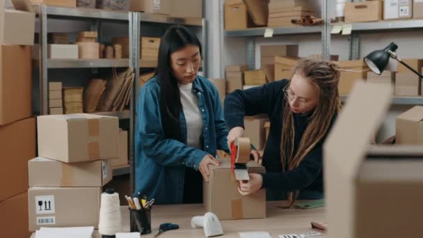 オンラインストアのオーナー。若い女性倉庫労働者は、自宅のオフィスで宅配小包ボックスを準備しながら、テープパッケージで小包を封印します。郵便サービスと小規模なビジネスコンセプト. — ストック動画