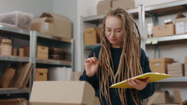 Спокойная женщина держит папку, записывая что-то на ней и готовясь отправить посылку клиенту. Девушка, работающая на складе. Концепция малого бизнеса. — стоковое видео