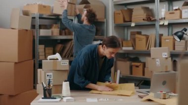 Ciddi bir beyaz adam masada duruyor ve erkek arkadaşı arka planda kutuları toplarken paketleri dikkatlice topluyor. Küçük işletme kavramı.