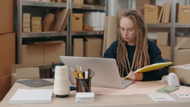 Beyaz tenli gözlüklü kadının dizüstü bilgisayarda oturduğu ve klavyede çalışırken paket detaylarını yazdığı belden yukarı bir görünüm. Posta servisi ve küçük işletme kavramı. — Stok video