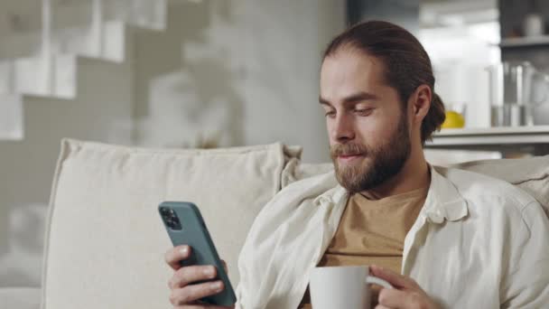 积极的男人坐在沙发上，拿着咖啡和智能手机 — 图库视频影像