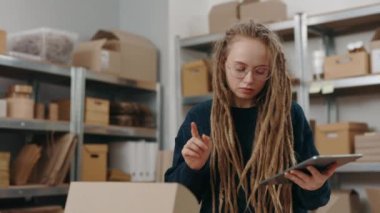 Sakin bir kadın tablet bilgisayarı kullanarak bir şeyleri karıştırıyor ve müşteriye paket göndermeye hazırlanıyor. Depoda çalışan kız. Küçük işletme kavramı.