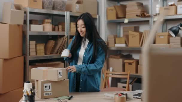 Midja upp porträtt bild av brunett asiatisk kvinna skanna paket medan du arbetar på lagret ensam. Hyllor med lådor i bakgrunden. Post service och småföretag koncept. — Stockvideo