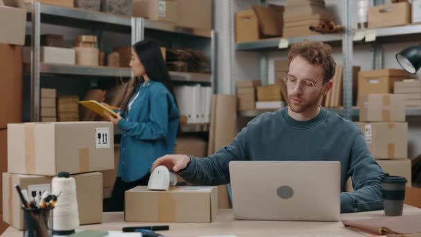Aandachtige blanke man met een bril op op de laptop en het scannen van pakketten terwijl zijn Aziatische brunette zijn pakketten sorteert. Concept voor kleine ondernemingen. — Stockvideo