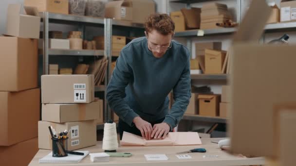 Midjan upp porträtt bild av ingefära uppmärksam man står vid bordet och förpackning paket med lätt leende på sin arbetsplats. Småföretagskoncept. — Stockvideo