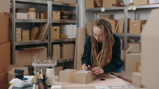 Caucasian female small business owner standing at the table and writing something at the cupboard box with felt tip pen while preparing parcel to the shipment. — Stock Video