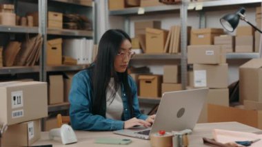 Özenli Asyalı kadın dizüstü bilgisayarın başında oturmuş ev ofisinde çalışırken klavyenin başına bir şeyler yazıyor. Posta servisi ve küçük işletme kavramı.